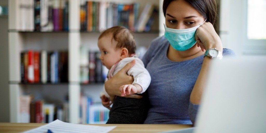 A young mother in a mask holds her baby while looking at her lap top in exhaustion
