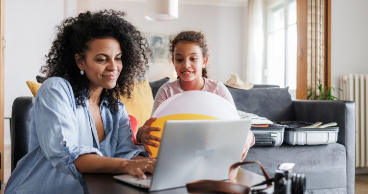 A mother works on her computer next to her daughter who is holding a beach ball.
