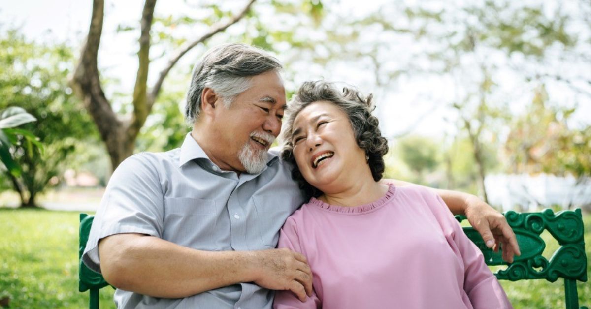 An older couple sit in the park while with big grins.