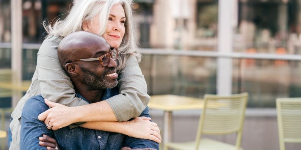 A woman hugs a man from behind with her arms draped around his shoulders