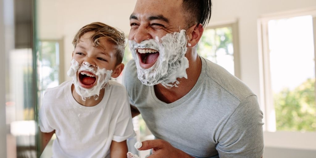 A young father and son smile in the mirror with shaving cream on their faces
